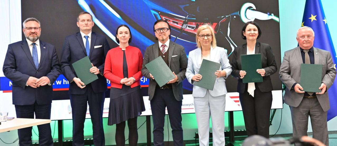 W podpisaniu umowy w dniu 13 maja 2026 r. udział wzięli (na zdjęciu od lewej): Mirosław Suchoń, Przewodniczący Komisji Infrastruktury, Cyprian Gronkiewicz, Prezes ElectroMobility Poland S.A., Eliza Zeidler, Sekretarz Stanu w Ministerstwie Aktywów Państwowych, Wojciech Balczun, Minister Aktywów Państwowych, Paulina Hennig-Kloska, Minister Klimatu i Środowiska, Dorota Zawadzka-Stępniak, Prezes Zarządu NFOŚiGW, Paweł Augustyn, Zastępca Prezesa Zarządu NFOŚiGW.